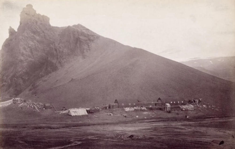 Dobová fotografie z roku 1883 zachycující umístění dřevěných budov a dispozice tábora pod vysokým černým tufovým vrcholem.