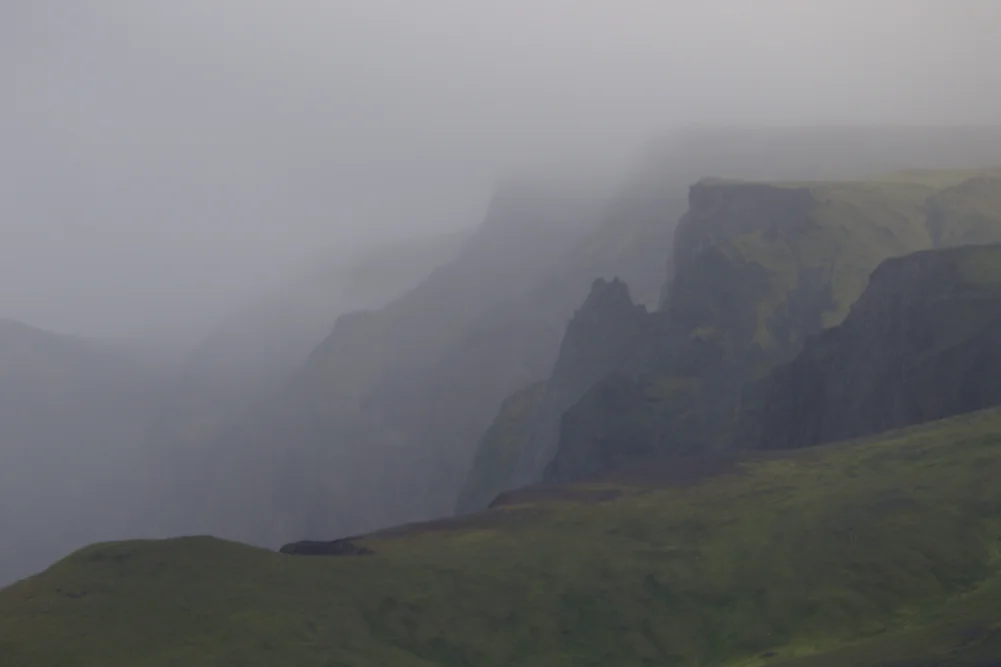 Strmé hory a skalní útesy pokryté zelenou vegetací obklopené mlhou a kondenzující vodou na ostrově Jan Mayen.